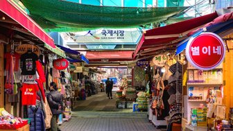 Ein belebter Markt mit bunten Ständen, die eine Vielzahl von Waren und Souvenirs anbieten, unter einem grünen Sonnenschutz.