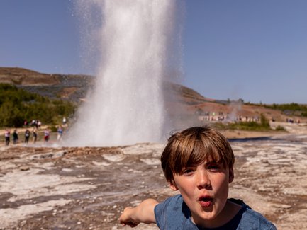 Ein Junge steht vor dem Geysir Strokkur, während er begeistert auf den sprudelnden Wasserstrahl zeigt.