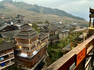 Blick auf die terrassierten Reisfelder und traditionellen Holzhäuser in Longsheng, umgeben von sanften Hügeln und Nebel.