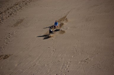 Zwei Personen rutschen fröhlich einen Sanddünenhang hinunter, während feiner Sand um sie herum aufwirbelt.