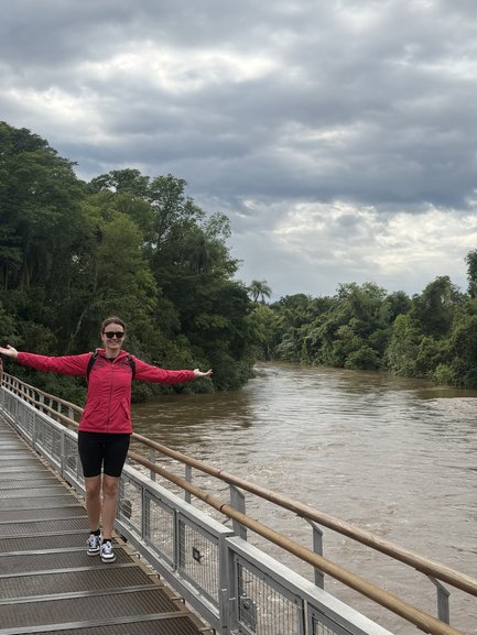 Eine Frau in einem pinken Kleid steht auf einer Holzplanke und breitet die Arme aus, um den malerischen Fluss zu genießen.