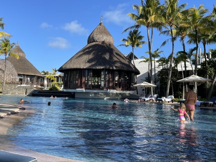 Ein tropischer Pool mit Palmen, umgeben von stilvollen Hütten, während Familienmitglieder im Wasser spielen und entspannen.