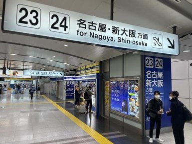 Ein belebter Bahnhof mit Schildern für die Züge nach Nagoya und Shin-Osaka, umgeben von Reisenden und Geschäften.