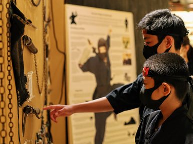 Zwei Kinder in Ninja-Kostümen betrachten mit Interesse eine Wand voller Ninja-Ausrüstung in einem Museum.
