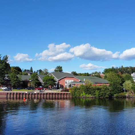 Ein ruhiger Fluss spiegelt die grüne Uferlandschaft und das Hotel Energie in Shawinigan unter einem strahlend blauen Himmel wider.