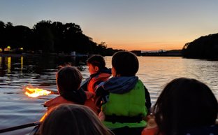 Eine Gruppe von Kindern in Kanus beobachtet den Sonnenuntergang, während sie mit Fackeln auf dem Wasser spielen.