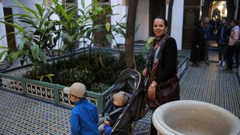 Eine Frau mit einem Kinderwagen steht in einem Innenhof, umgeben von üppigen Pflanzen und bunten Fliesen.