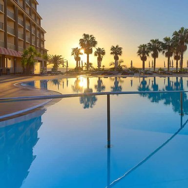 Ein atemberaubender Sonnenuntergang spiegelt sich im glitzernden Pool eines Hotels, umgeben von Palmen und Liegen.