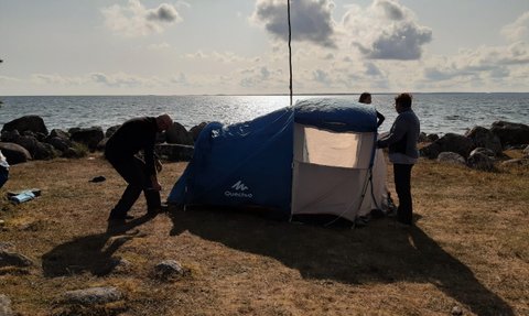 Drei Personen bauen ein Zelt am Ufer eines Sees auf, während die Sonne am Horizont scheint und Wolken am Himmel schweben.