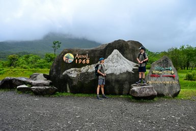Zwei Wanderer stehen vor einer großen Gesteinsmarkierung im Arenal-Nationalpark, umgeben von üppiger grüner Natur.