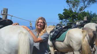 Eine fröhliche Frau lächelt, während sie ein Pferd streichelt, umgeben von weiteren gesattelten Pferden im Hintergrund.