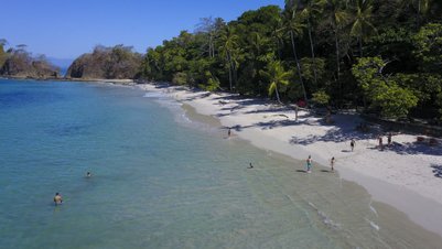 Ein sonniger Strand mit klarem Wasser, wo Menschen im Wasser schwimmen und am Ufer spazieren gehen.