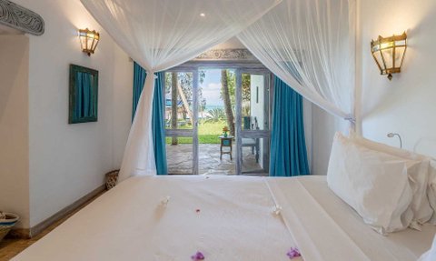 Ein helles Zimmer mit einem großen Bett, umgeben von einem Moskitonetz, bietet einen Blick auf den Strand durch große Fenster.