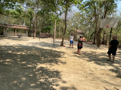 Eine Gruppe von Menschen spielt Basketball in einem schattigen, sandigen Bereich mit Bäumen im Hintergrund.
