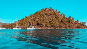 Drei Segelboote liegen ruhig im glitzernden Wasser, umgeben von einer bewaldeten Insel unter strahlend blauem Himmel.