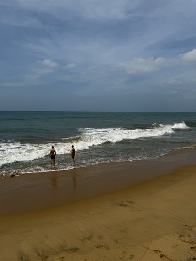 Zwei Personen stehen am Strand und beobachten die Wellen, während der Himmel leicht bewölkt ist und das Wasser glitzert.