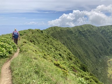 Ein Wanderer mit einem Rucksack geht einen schmalen Pfad entlang, umgeben von üppigem Grün und steilen Hängen.