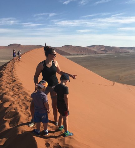 Eine Frau steht auf einer Sanddüne und zeigt auf die Landschaft, während zwei Kinder aufmerksam zu ihr schauen.