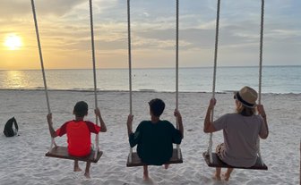 Drei Personen schwingen entspannt am Strand, während die Sonne langsam am Horizont untergeht und den Himmel in warmen Farben färbt.