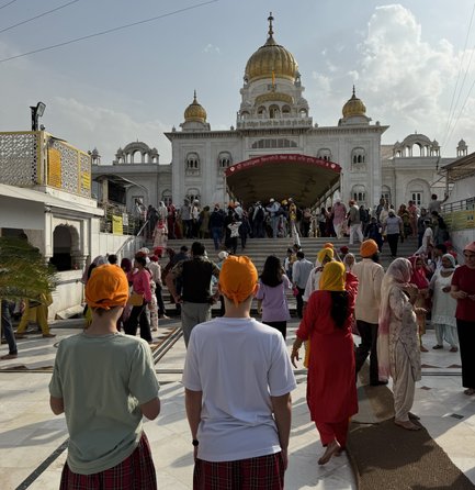 Zwei Personen mit orangefarbenen Turbanen stehen vor dem Gurudwara Sri Bangla Sahib, umgeben von einer lebhaften Menschenmenge.