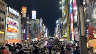 Die belebte Dotonbori-Straße in Osaka ist nachts erleuchtet, mit Menschenmengen, die am Fluss entlang spazieren.