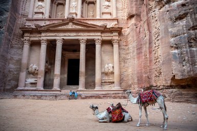 Zwei Kamele mit bunten Sätteln ruhen im Sand vor der beeindruckenden Fassade eines antiken Gebäudes in Petra.