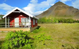 Ein rotes Ferienhaus mit einer Veranda steht vor einem majestätischen Berg, umgeben von grünen Wiesen und blühenden Pflanzen.