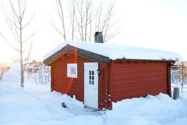Eine rote Holzhütte steht in einer verschneiten Landschaft, umgeben von kahlen Bäumen und sanften Hügeln.