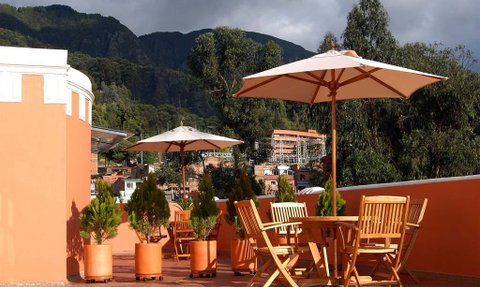 Eine sonnige Terrasse mit Holzmöbeln und Sonnenschirmen, umgeben von grünen Pflanzen und einer beeindruckenden Berglandschaft.