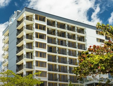 Modernes Hotelgebäude mit vielen Balkonen, umgeben von Bäumen und einem strahlend blauen Himmel.
