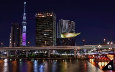 Die Tokyo Skytree leuchtet in bunten Farben, während moderne Gebäude und eine Brücke den nächtlichen Fluss säumen.