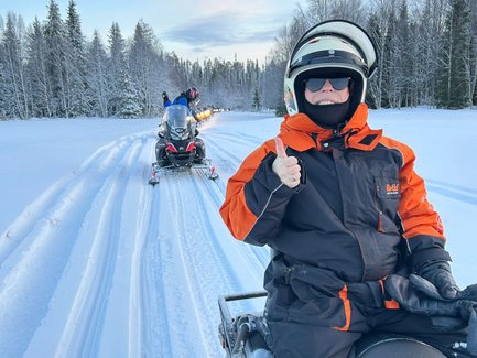 Eine Person in warmer Winterkleidung gibt ein Daumen-hoch-Zeichen, während sie auf einem Schneemobil fährt.