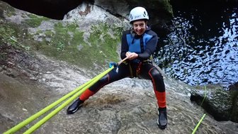 Eine Person in einem Neoprenanzug klettert an einer steilen Felswand mit einem Seil, umgeben von grünem Moos und Wasser.