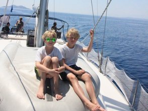 Zwei Jungen mit blonden Haaren sitzen auf dem Bug eines Segelbootes, umgeben von glitzerndem Wasser und klarem Himmel.