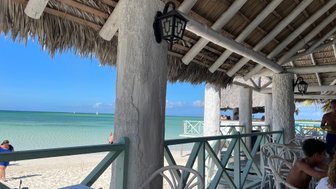 Ein Blick auf einen Strand mit klarem Wasser, umgeben von einem schattigen Restaurant mit Strohdach und weißen Säulen.