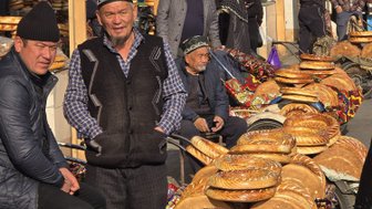 Auf einem Markt stehen mehrere Männer neben einem Wagen voller frisch gebackenen Broten, umgeben von anderen Käufern.