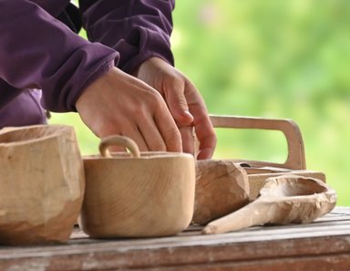Eine Hand formt sorgfältig ein Holzobjekt, während mehrere geschnitzte Holzgegenstände auf einem Tisch liegen.