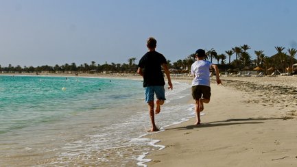 Zwei Kinder rennen fröhlich am Strand entlang, während die Wellen sanft an den Ufer schlagen und die Sonne scheint.