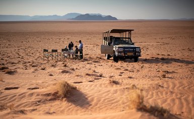 Unter einem klaren Himmel genießen vier Personen ein Frühstückstisch im Sand, umgeben von der majestätischen Wüstenlandschaft.