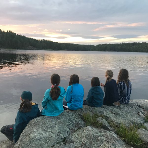 Sechs Kinder sitzen auf einem Felsen am Ufer eines ruhigen Sees und beobachten den Sonnenuntergang über den Bäumen.