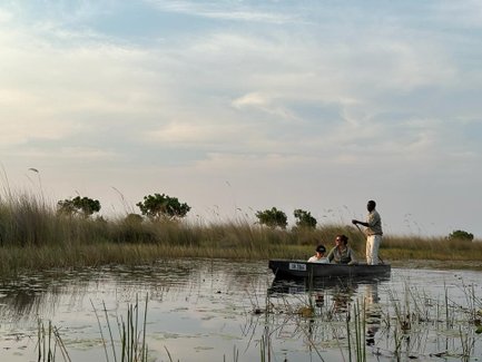 Eine Gruppe von drei Personen fährt in einem Boot durch eine ruhige Wasserlandschaft mit hohem Gras und Bäumen im Hintergrund.