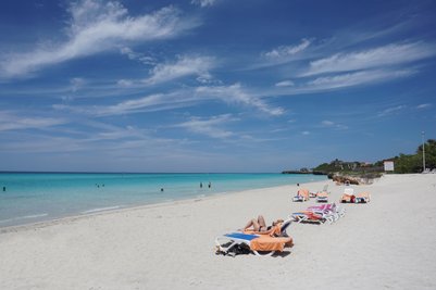 Ein sonniger Strand mit weißen Sandflächen, Liegen und Menschen, die im klaren Wasser schwimmen und sich entspannen.