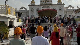 Zwei Personen mit orangefarbenen Turbanen stehen vor dem Gurudwara Sri Bangla Sahib, umgeben von einer lebhaften Menschenmenge.