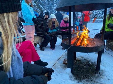 Eine Gruppe von Menschen sitzt um ein Feuer im Schnee, während sie Würstchen grillen und die winterliche Landschaft genießen.