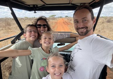 Eine fröhliche Familie posiert in einem offenen Fahrzeug auf einer roten Piste, umgeben von trockener Savanne.