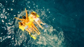 Vier Personen schwimmen fröhlich im klaren blauen Wasser, während Sonnenstrahlen auf die Oberfläche scheinen.