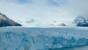 Ein majestätischer Gletscher erstreckt sich über das Wasser, umgeben von schneebedeckten Bergen und einem klaren blauen Himmel.