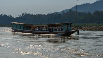 Ein traditionelles Boot fährt auf einem ruhigen Fluss, umgeben von üppiger Vegetation und sanften Hügeln im Hintergrund.