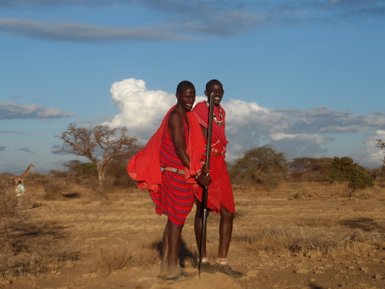 Zwei fröhliche Maasai-Krieger in leuchtend roten Gewändern posieren in der afrikanischen Savanne unter einem blauen Himmel.