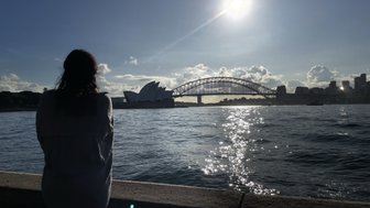 Eine Person sitzt am Ufer und betrachtet die Abendsonne über dem Hafen, mit der Oper im Vordergrund.